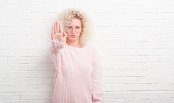 Young blonde woman with curly hair over white brick wall doing stop sing with palm of the hand. Warning expression with negative and serious gesture on the face. - Photo, Image