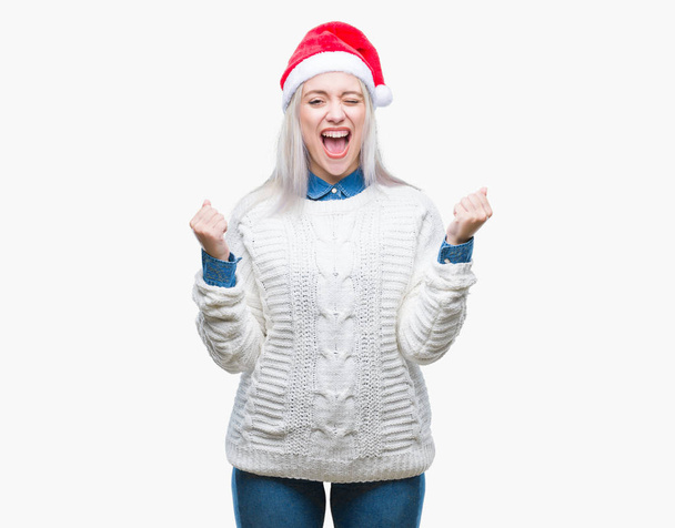 Young blonde woman wearing christmas hat over isolated background celebrating surprised and amazed for success with arms raised and open eyes. Winner concept. - Foto, imagen