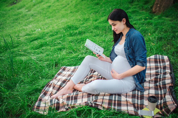 Portrait of a happy black hair and proud pregnant woman in the park. The female model is sitting on grass and The female model is reading a book - 写真・画像