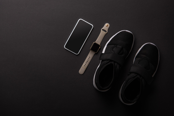 bovenaanzicht gearrangeerde smartphone, smartwatch of sneakers geïsoleerd op zwart, minimalistische concept  - Foto, afbeelding