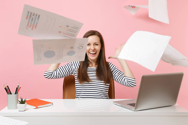 Smějící se dívka zvracel papírové dokumenty při práci na projektu, sedí v kanceláři s notebookem před víkendem izolované na růžovém pozadí. Úspěch obchodní kariéru koncept. Kopírovat prostor - Fotografie, Obrázek