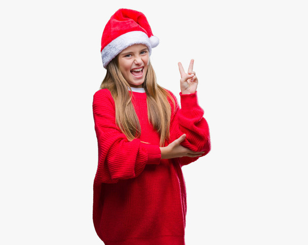 Όμορφη κοπέλα φορώντας καπέλο Χριστουγέννων πέρα από το απομονωμένο υπόβαθρο χαμογελώντας με χαρούμενο πρόσωπο που κλείνει το μάτι στην κάμερα κάνει το σήμα της νίκης. Αριθμός δύο. - Φωτογραφία, εικόνα