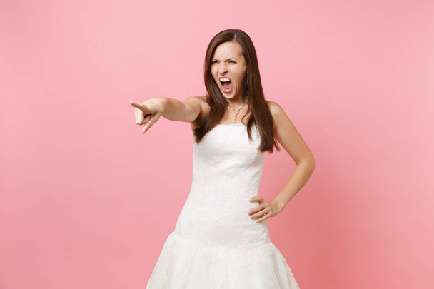 Porträt einer wütend irritierten Braut im weißen Hochzeitskleid, die schreiend mit dem Zeigefinger zur Seite zeigt, isoliert auf rosa Hintergrund. Hochzeitsfeier Konzept. Kopierfläche für Werbung - Foto, Bild
