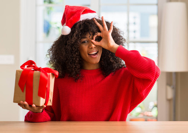 Mutlu yüz el parmaklarının arasından seyir göz üzerinde ok işaretiyle yapıyor gülümseyen ile mevcut giyen Noel kırmızı şapka tutan Afrikalı-Amerikalı kadın - Fotoğraf, Görsel