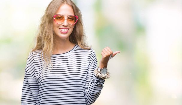 Beautiful young blonde woman wearing sunglasses over isolated background smiling with happy face looking and pointing to the side with thumb up. - Foto, Bild