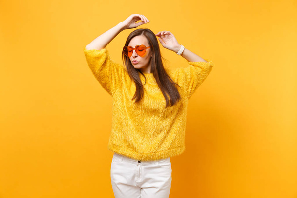 Portrait of relaxed young woman in fur sweater, white pants and heart orange glasses rising hands up isolated on bright yellow background. People sincere emotions, lifestyle concept. Advertising area - Foto, afbeelding