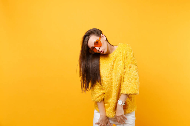 Retrato de una joven muy divertida en suéter de piel pantalones blancos gafas corazón naranja soplando labios aislados sobre fondo amarillo brillante. Gente emociones sinceras, concepto de estilo de vida. Área publicitaria
 - Foto, imagen