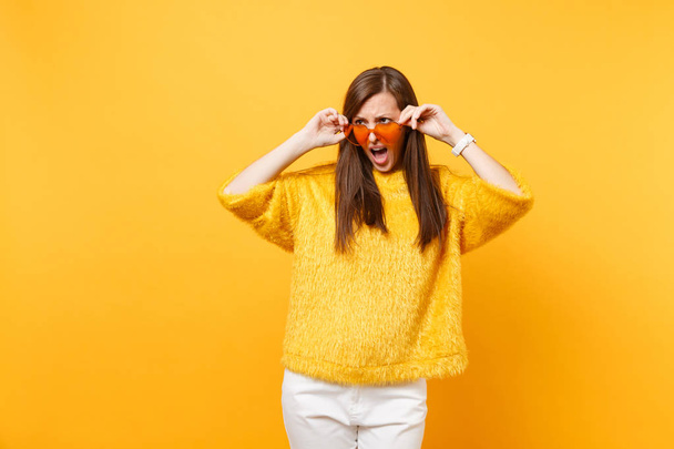 Upset offended young woman in fur sweater, white pants holding heart orange glasses looking aside isolated on bright yellow background. People sincere emotions, lifestyle concept. Advertising area - Фото, изображение