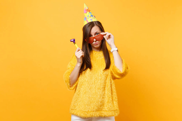 Verwirrte Frau mit Geburtstagsmütze und orangefarbener lustiger Brille mit Pfeife, die isoliert auf gelbem Hintergrund feiert. Menschen aufrichtige Emotionen Lifestyle-Konzept. Werbefläche - Foto, Bild