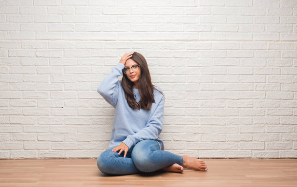 Young brunette woman sitting on the floor over white brick wall stressed with hand on head, shocked with shame and surprise face, angry and frustrated. Fear and upset for mistake. - 写真・画像