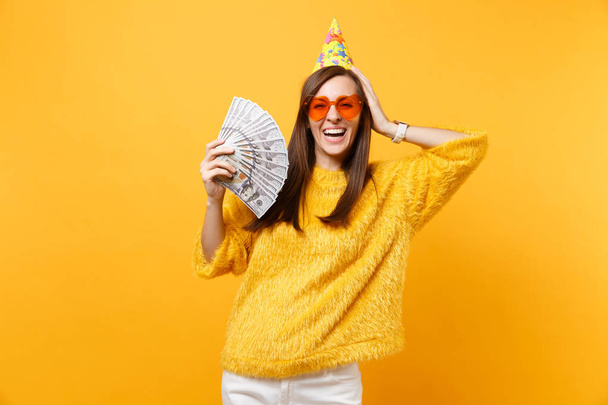 Mujer joven riendo en gafas de corazón naranja sombrero de cumpleaños poniendo la mano en la cabeza sosteniendo paquete un montón de dólares dinero en efectivo, celebrando aislado en el fondo amarillo. Personas emociones sinceras, estilo de vida
 - Foto, imagen