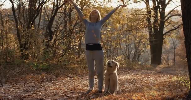 Młoda kobieta z jej pies pies myśliwski podrzucanie liści nawet w jesień las park - Materiał filmowy, wideo