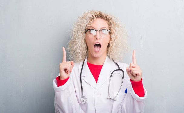 junge blonde Ärztin über grunge graue Wand erstaunt und überrascht Blick nach oben und zeigt mit Fingern und erhobenen Armen. - Foto, Bild