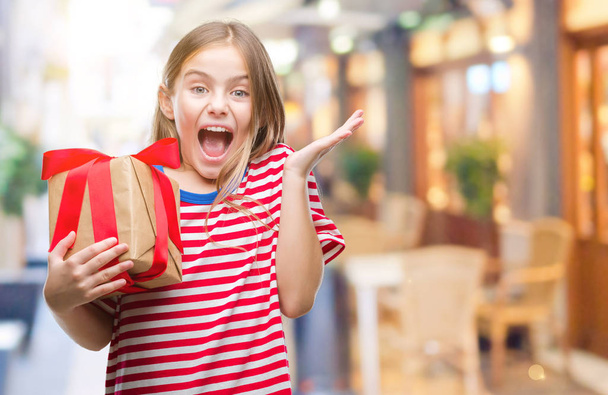 Young beautiful girl giving christmas or valentine gift over isolated background very happy and excited, winner expression celebrating victory screaming with big smile and raised hands - Foto, Imagen