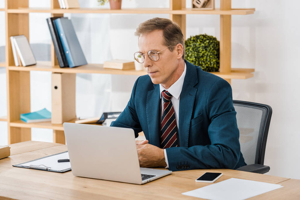adult serious businessman in glasses sitting at table and using laptop in office - Φωτογραφία, εικόνα