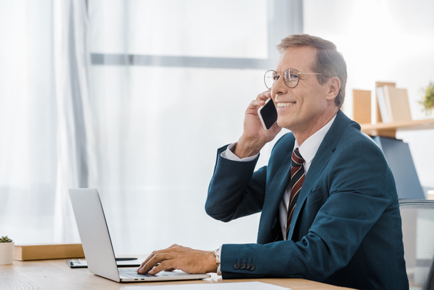 adulto sorridente empresário em óculos falando no smartphone e usando laptop no escritório
 - Foto, Imagem
