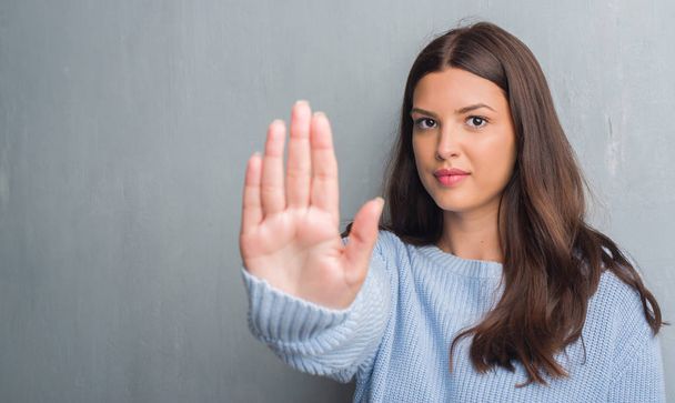 Jonge brunette vrouw over grunge grijs muur met open hand doen stopbord met ernstige en vol vertrouwen meningsuiting, defensie gebaar - Foto, afbeelding