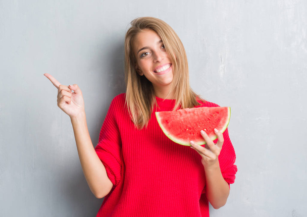 Beautiful young woman over grunge grey wall eating water melon very happy pointing with hand and finger to the side - Photo, Image