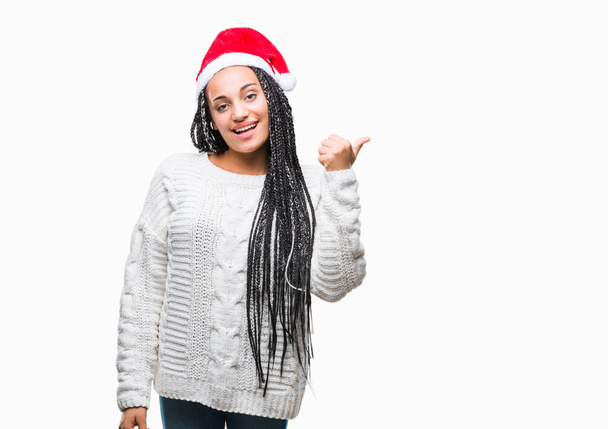 ヤング編見て、親指側に上向きの幸せそうな顔を浮かべて隔離された背景にクリスマスの帽子をかぶって髪アフリカ系アメリカ人の女の子. - 写真・画像