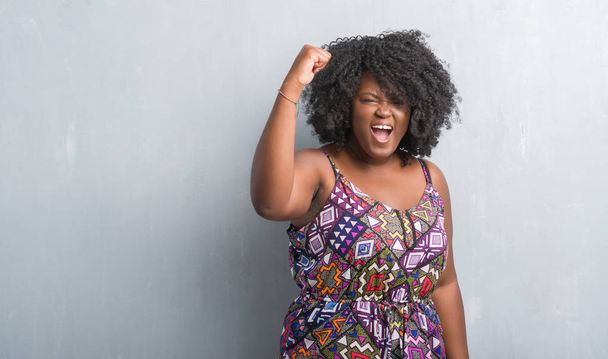 Młody african american kobieta szary mur sobie kolorowy strój zły i szalony, podnosząc pięść, sfrustrowani i wściekły krzykiem z gniewu. Wściekłość i agresywne koncepcja. - Zdjęcie, obraz