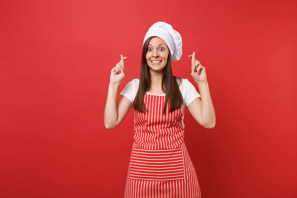 Housewife female chef cook or baker in striped apron, white t-shirt, toque chefs hat isolated on red wall background. Happy woman keeping fingers crossed and making wish. Mock up copy space concept - Фото, изображение