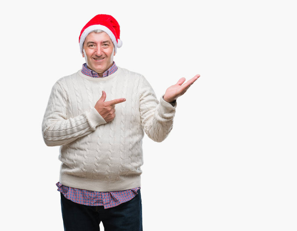 bel homme âgé portant chapeau de Noël sur fond isolé étonné et souriant à la caméra tout en présentant avec la main et pointant du doigt
. - Photo, image