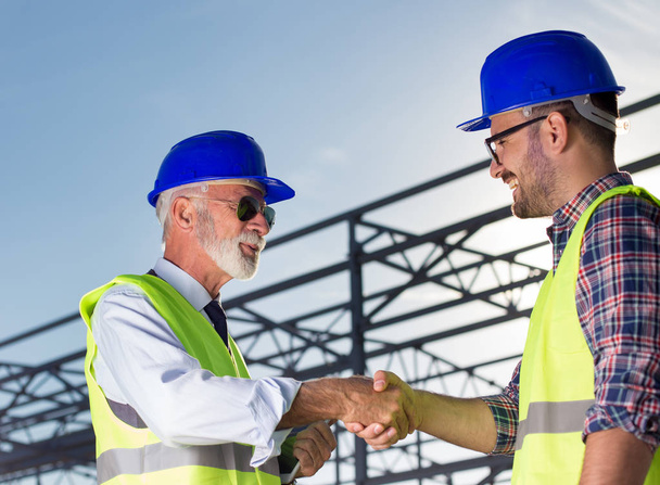 Два инженера пожимают руку на строительной площадке с металлической конструкцией на заднем плане
 - Фото, изображение