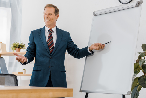 Lächelnder Geschäftsmann im Anzug zeigt auf weiße Tafel im Büro - Foto, Bild