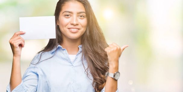 Giovane donna araba in possesso di carta bianca su sfondo isolato che punta e mostra con il pollice verso l'alto sul lato con il viso felice sorridente
 - Foto, immagini