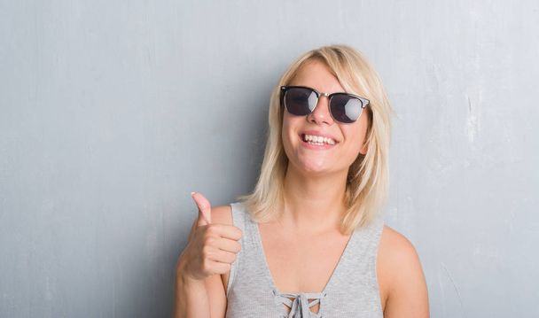 Adulto mulher caucasiana sobre grunge parede cinza usando óculos de sol feliz com grande sorriso fazendo sinal ok, polegar com os dedos, excelente sinal
 - Foto, Imagem