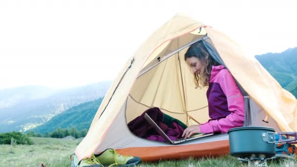 Happy tourist woman uses laptop in a tent, freelancer prints in nature in a journey against the backdrop of a mountain landscape. - Footage, Video