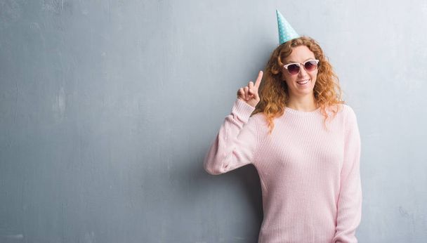 Jeune femme rousse sur un mur gris grunge portant un chapeau d'anniversaire surpris par une idée ou une question pointant du doigt avec un visage heureux, numéro un
 - Photo, image