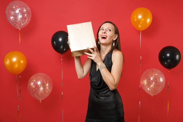 Happy young girl in little black dress celebrating holding golden box with gift present on bright red background air balloons. St. Valentine's Day Happy New Year birthday mockup holiday party concept - Photo, Image