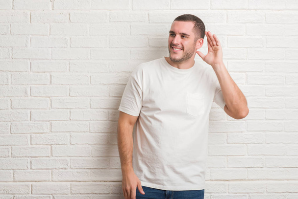 Kavkazské mladík stojící nad bílou cihlová zeď s úsměvem s rukou přes ucho naslouchá slyšení zvěsti nebo drby. Hluchota koncept. - Fotografie, Obrázek