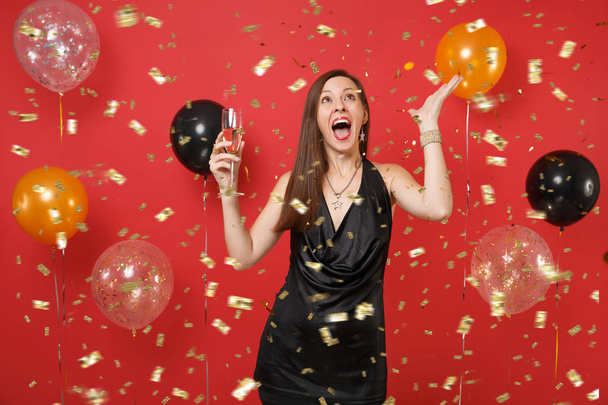 リトル ブラック ドレスを祝って、シャンパン グラスを持って、明るい赤の背景空気風船に紙吹雪をキャッチで若い女性を興奮させた。幸せな新年、誕生日モックアップ休日党概念 - 写真・画像