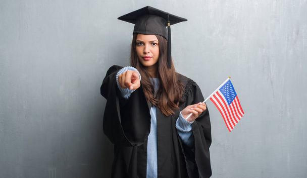 Jeune femme brune sur le mur gris grunge portant uniforme diplômé tenant drapeau de l'Amérique pointant du doigt à la caméra et à vous, signe de la main, geste positif et confiant de l'avant
 - Photo, image