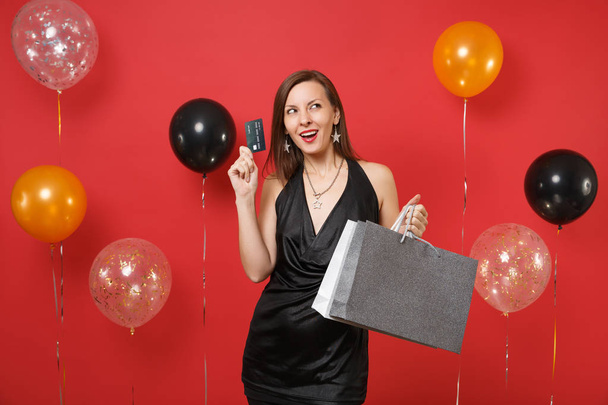 Curious young girl in black dress holding credit card, colored packages bags with purchases after shopping on bright red background air balloons. Happy New Year, birthday mockup holiday party concept - Foto, afbeelding