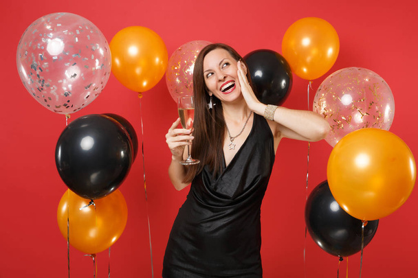 Dreamy girl in black dress celebrating looking up hold glass of champagne, keeping hand on face on bright red background air balloons. Women's Day Happy New Year birthday mockup holiday party concept - Photo, Image