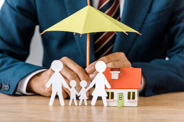 Male hands with paper cut family, house model and umbrella on wooden table, life insurance - Photo, Image