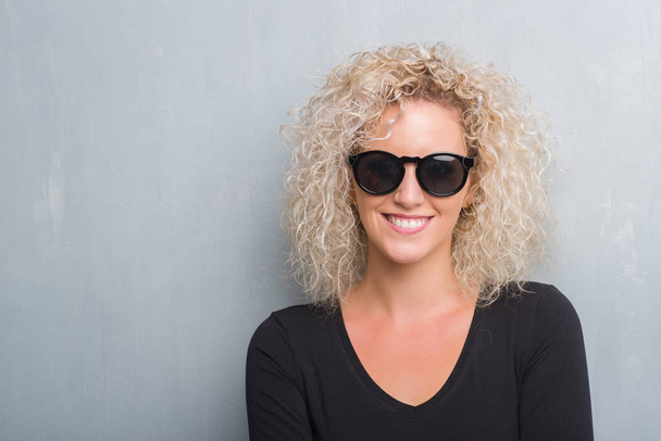Young blonde woman with curly hair over grunge grey background happy face smiling with crossed arms looking at the camera. Positive person. - 写真・画像