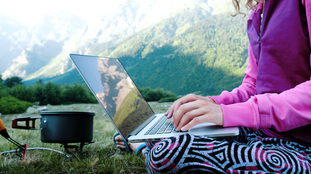 Tourist woman freelancer uses a computer in nature and sits on the grass next to a gas torch. Mountains are reflected in the laptop screen - Footage, Video