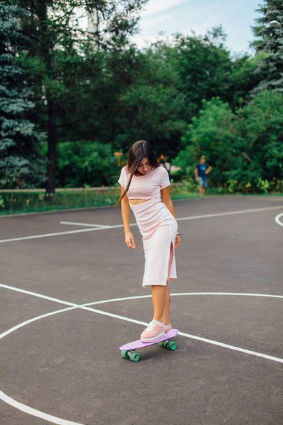 Portrait of a smiling charming brunette female standing on her skateboard on a basketball court. - Photo, Image