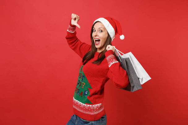 Excited Santa girl keeping mouth wide open pointing thumb on herself hold packages bags with purchases after shopping isolated on red background. Happy New Year 2019 celebration holiday party concept - Photo, Image