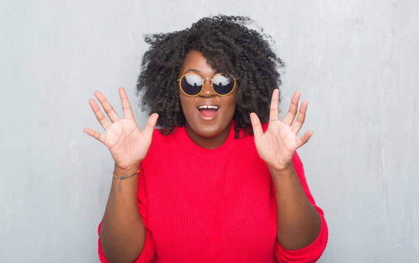 Joven afroamericana más tamaño mujer sobre gris grunge pared usando gafas de sol retro celebrando loco y sorprendido por el éxito con los brazos levantados y los ojos abiertos gritando emocionado. Concepto ganador
 - Foto, imagen