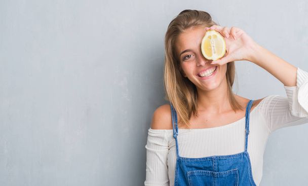 Bella giovane donna sopra il muro grigio grunge che tiene un limone con un viso felice in piedi e sorridente con un sorriso fiducioso che mostra i denti
 - Foto, immagini
