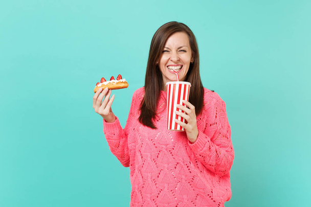 Giovane donna sorridente in maglia rosa maglione tenuta in mano eclair torta, tazza di plastica di cola o soda isolata su sfondo blu parete, studio ritratto. Concetto di stile di vita. Falsificare spazio di copia
 - Foto, immagini