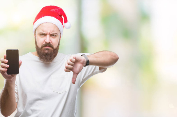 Young caucasian man wearing christmas hat showing smartphone screen over isolated background with angry face, negative sign showing dislike with thumbs down, rejection concept - Фото, изображение