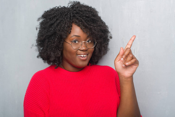 Jonge african american vrouw over grijs grunge muur dragen winter trui en glazen vinger omhoog met succesvolle idee. Verlaten en gelukkig. Nummer één. - Foto, afbeelding