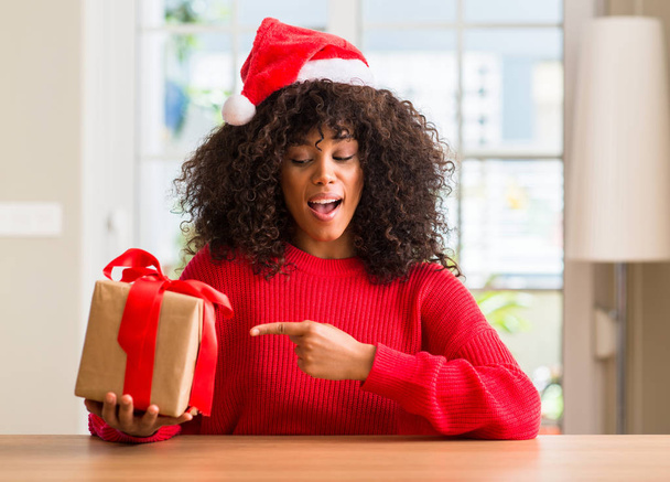 Femme afro-américaine tenant présent portant chapeau rouge de Noël très heureux pointant avec la main et le doigt
 - Photo, image
