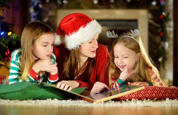 Young mother and her two little daughters reading a book by a Christmas tree in cozy living room in winter - Fotó, kép